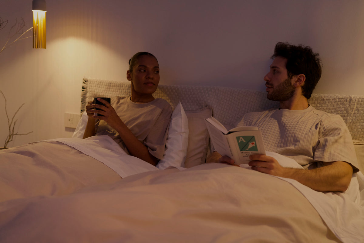 A couple in bed in the evening, one reading, one holding a phone, under a warm bedside lamp